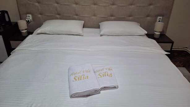 A neatly made bed with two pillows, a nightstand with a lamp, and two towels with text on them. - Hotel Vila Silia (Skopje)