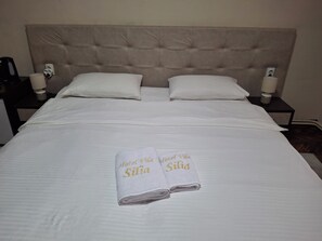 Een net opgemaakt bed met twee kussens, een nachtkastje met een lamp, en twee handdoeken met tekst erop.