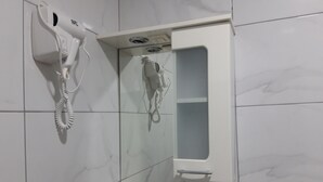 A bathroom with a white hairdryer mounted on the wall, a mirror, and a small shelf. - Hotel Vila Silia (Skopje)
