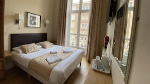 A hotel room with a large bed, a bedside table, a mirror, a window with curtains, and a view of buildings outside. - Belvedere Hotel (London)