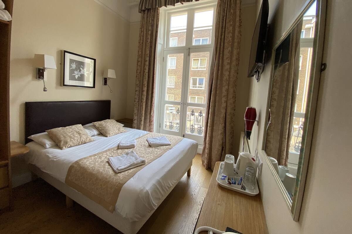 A hotel room with a large bed, a bedside table, a mirror, a window with curtains, and a view of buildings outside.