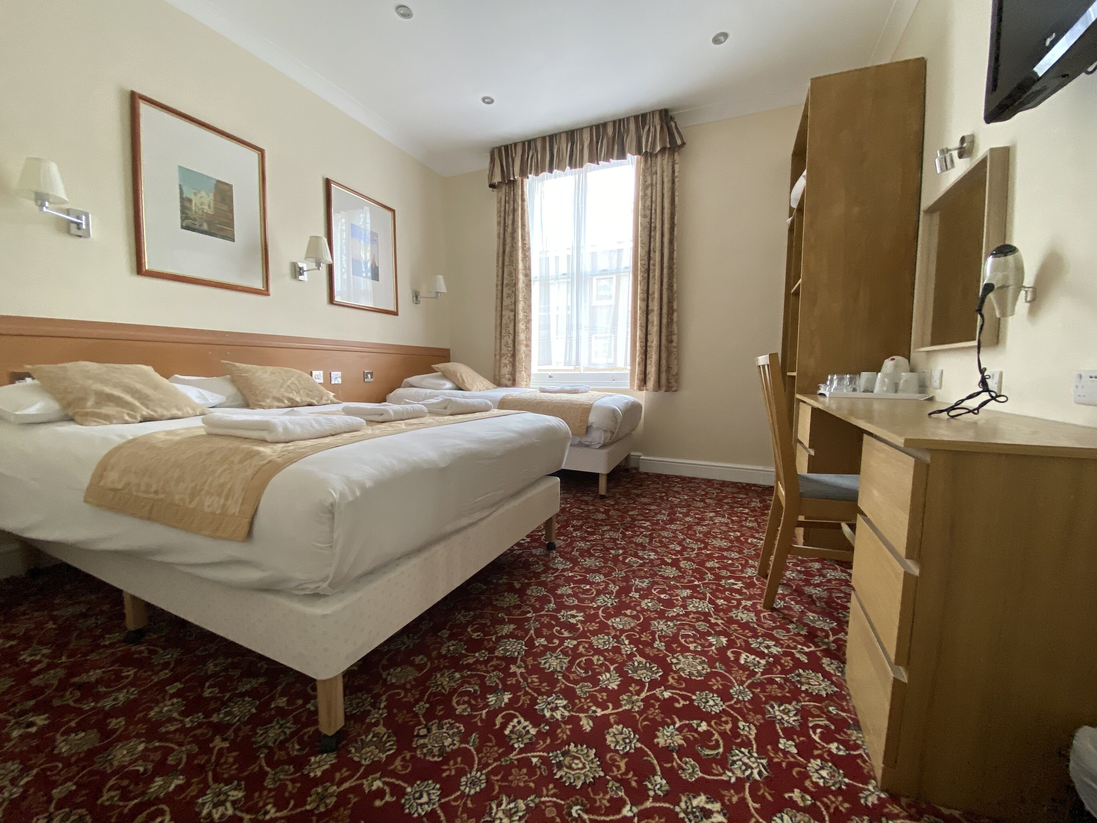 A hotel room with two beds, a wooden wardrobe, a desk with a mirror, and a window with curtains.