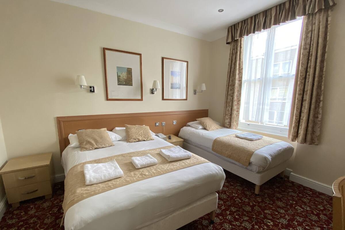 A hotel room with two beds, a nightstand, a window with curtains, and two framed pictures on the wall.