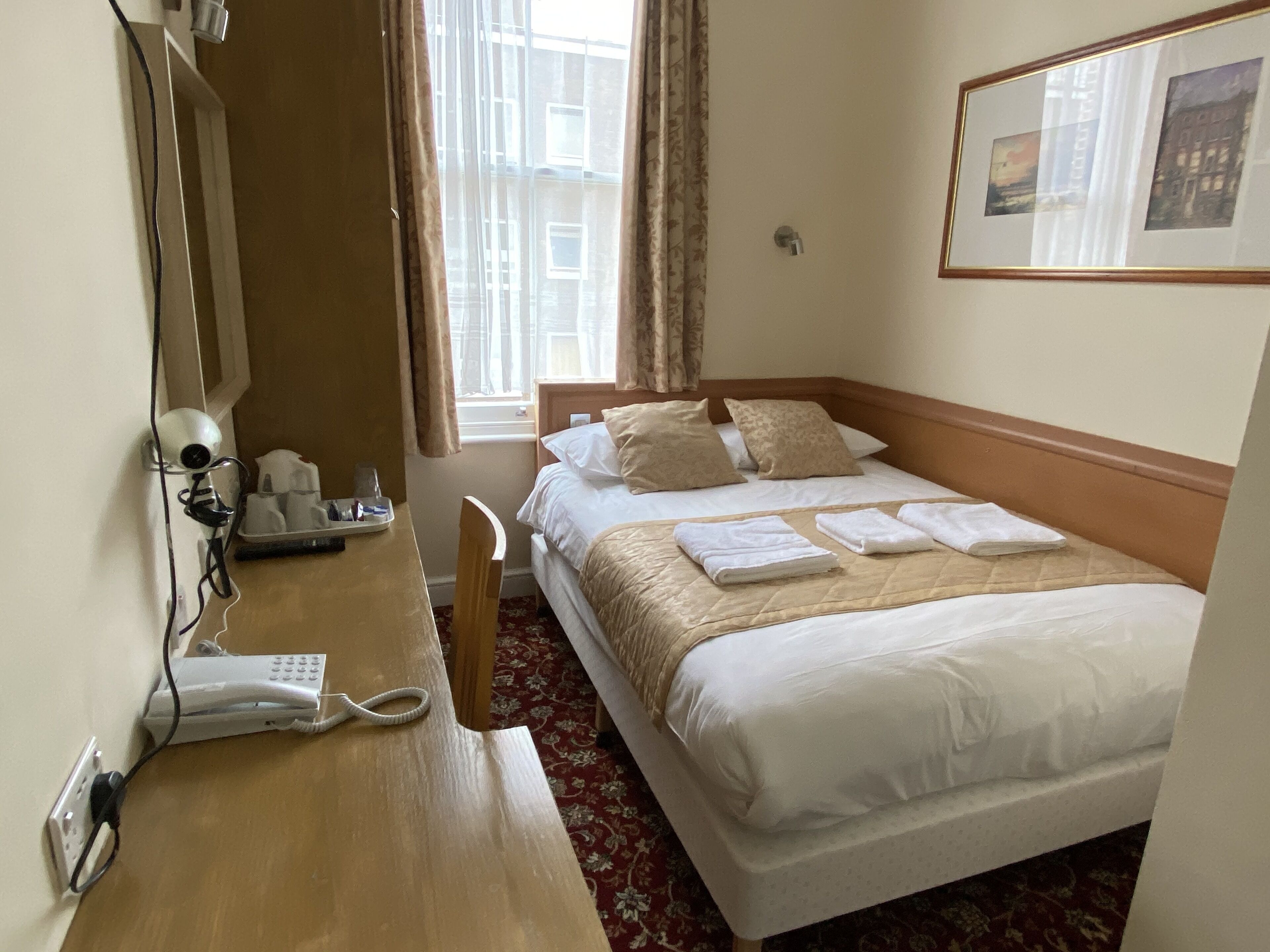 A hotel room with a bed, a desk with a telephone, a chair, and a window with curtains.