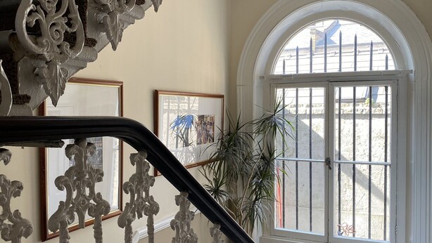 A grand staircase with ornate white railings and a black handrail, leading to a room with a large arched window and a potted plant. - Belvedere Hotel (London)