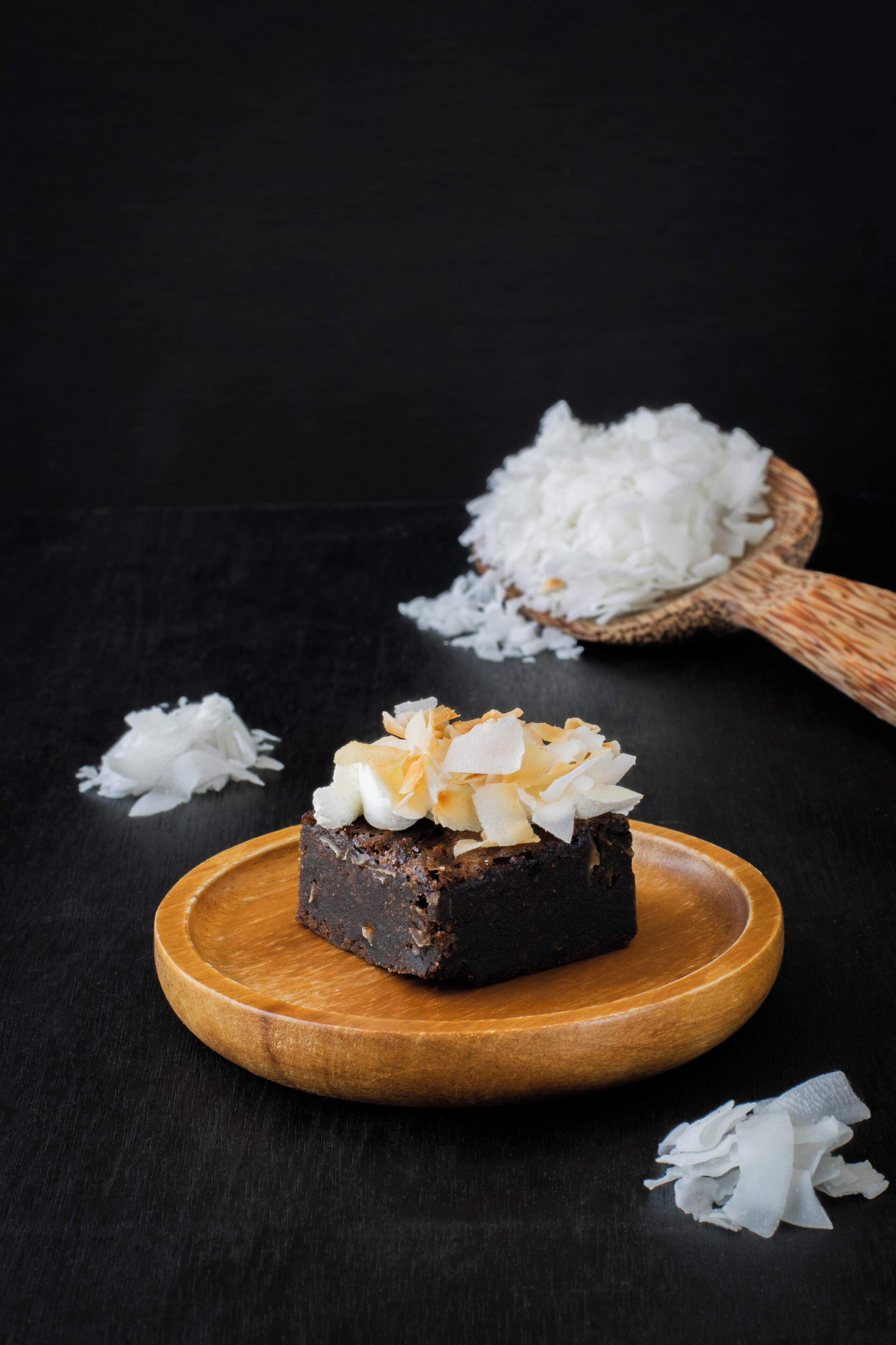Une part de gâteau au chocolat sur une assiette en bois, recouverte de copeaux de noix de coco, le tout posé sur une surface sombre et entouré de râpure de noix de coco.