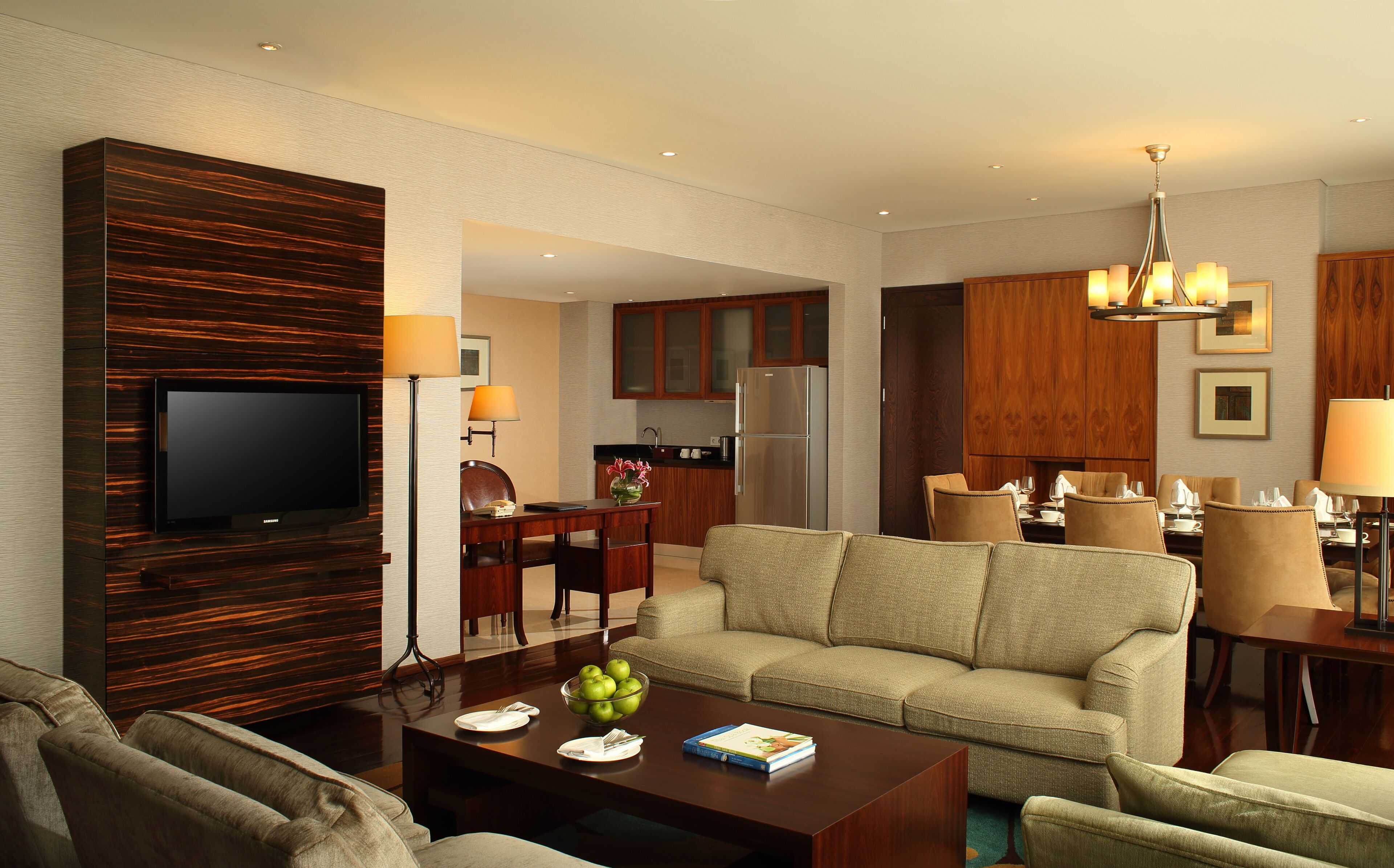 A modern hotel room with a flat-screen TV, a sofa, a dining area with chairs, a kitchen with wooden cabinets, and a chandelier.