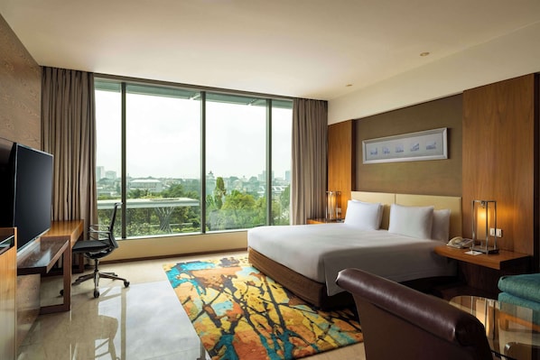 A hotel room with a large bed, a desk with a TV, a chair, a bedside table with a lamp, and a view of the city through large windows. - Hilton Bandung (Bandung)