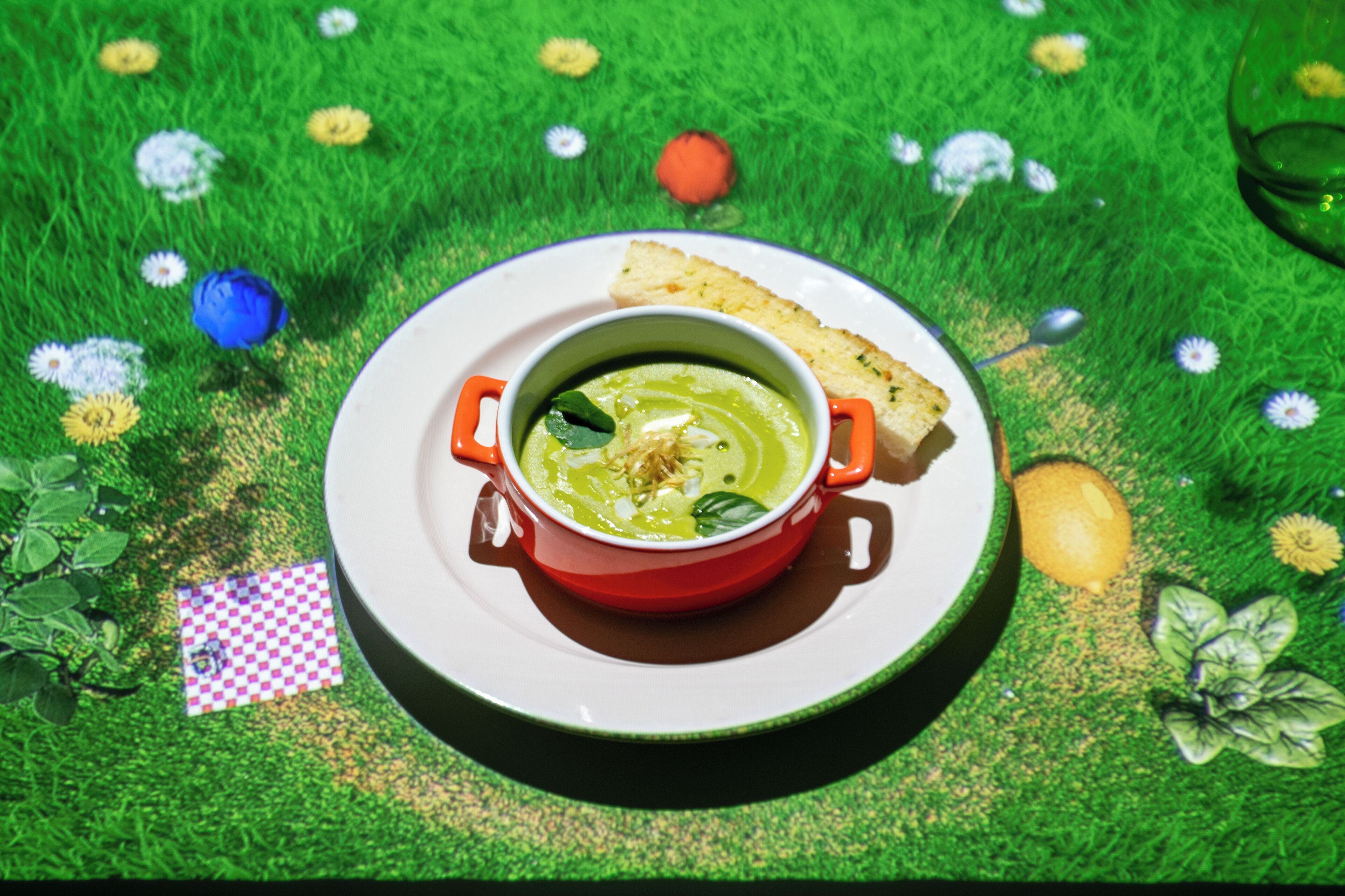 A bowl of soup with bread on a plate, set on a green surface with flowers.