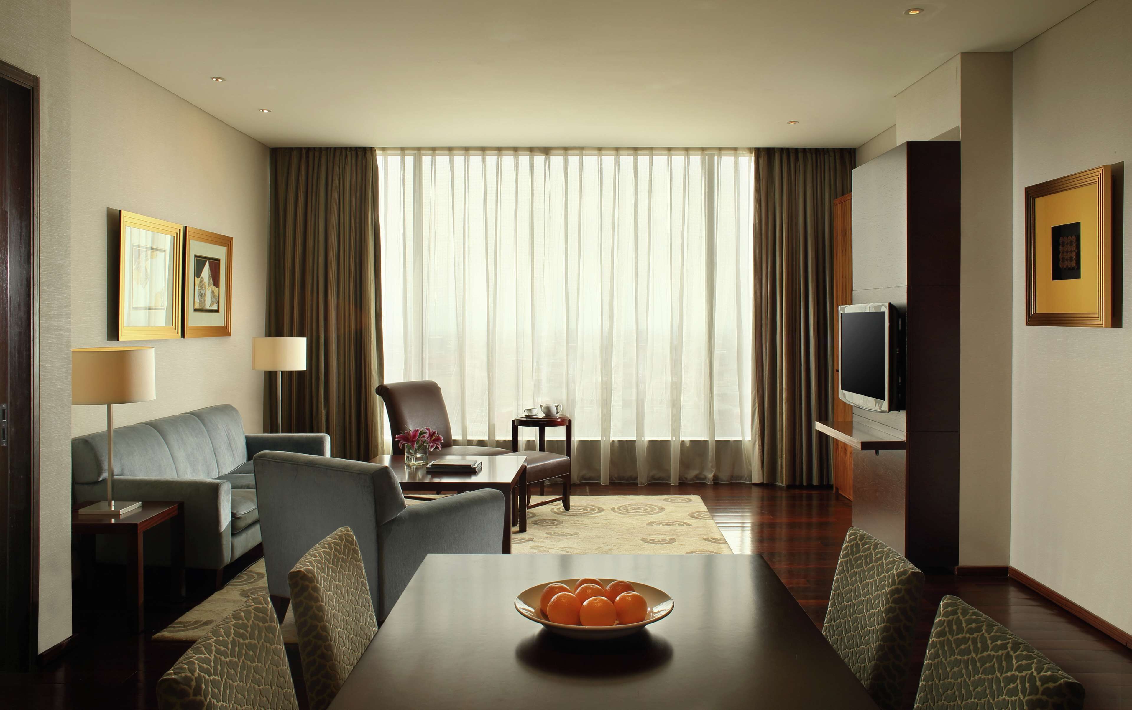 Een hotelkamer met een eettafel, stoelen, een televisie, een bank, een bijzettafeltje en een raam met gordijnen.