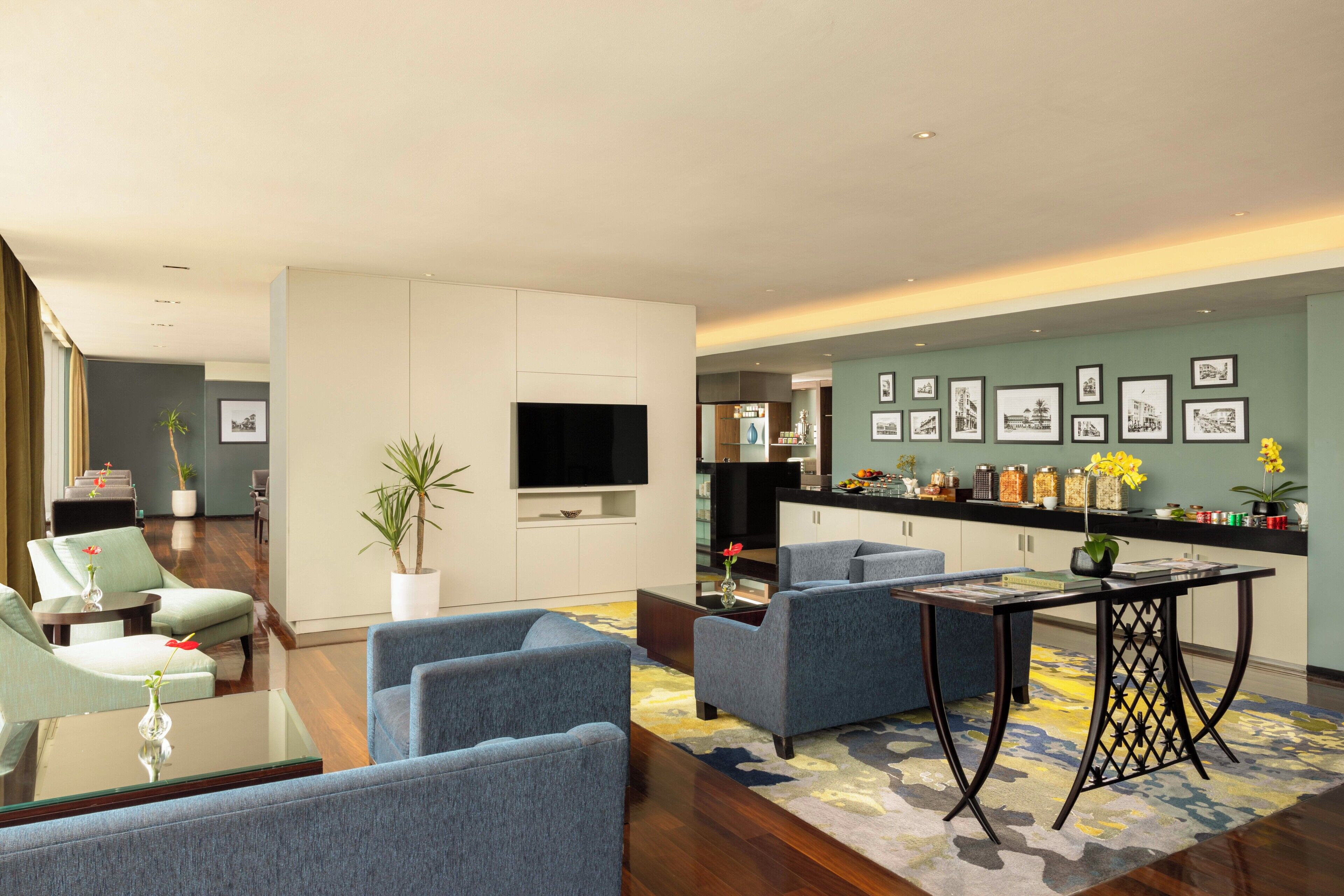 Una sala moderna con televisor de pantalla plana, sofás azules, una mesa de centro y una zona de cocina con refrigerador y varios utensilios.
