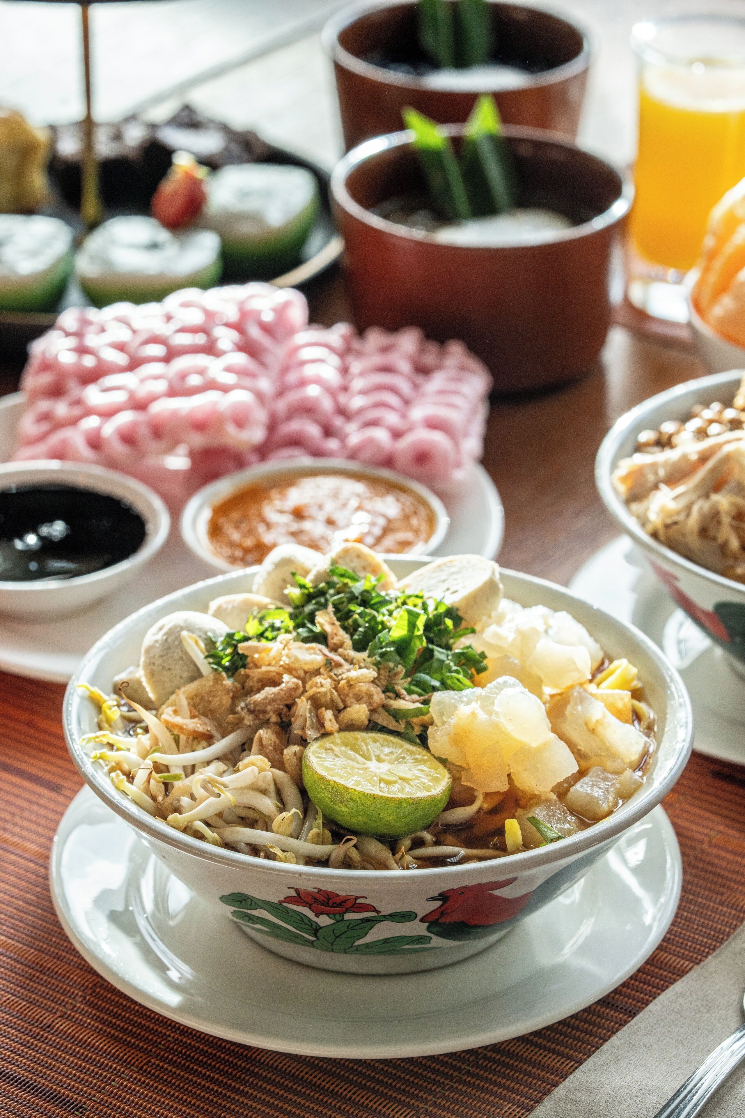 A bowl of noodle soup with lime and herbs, surrounded by various condiments and dishes on a table.