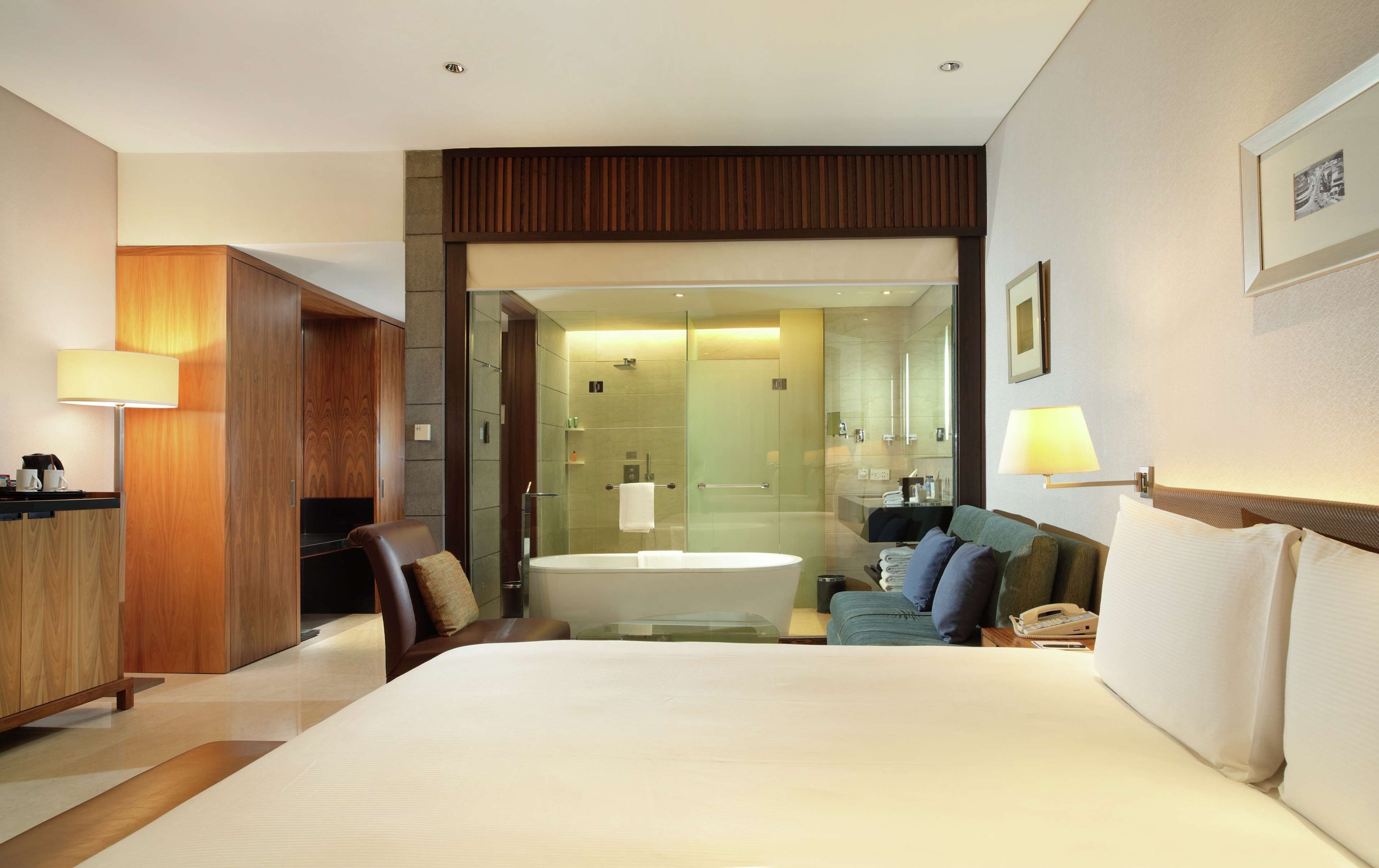 A modern hotel room with a large bed, a sitting area with a sofa and armchairs, a desk with a lamp, a bathroom with a bathtub and shower, and a large mirror.
