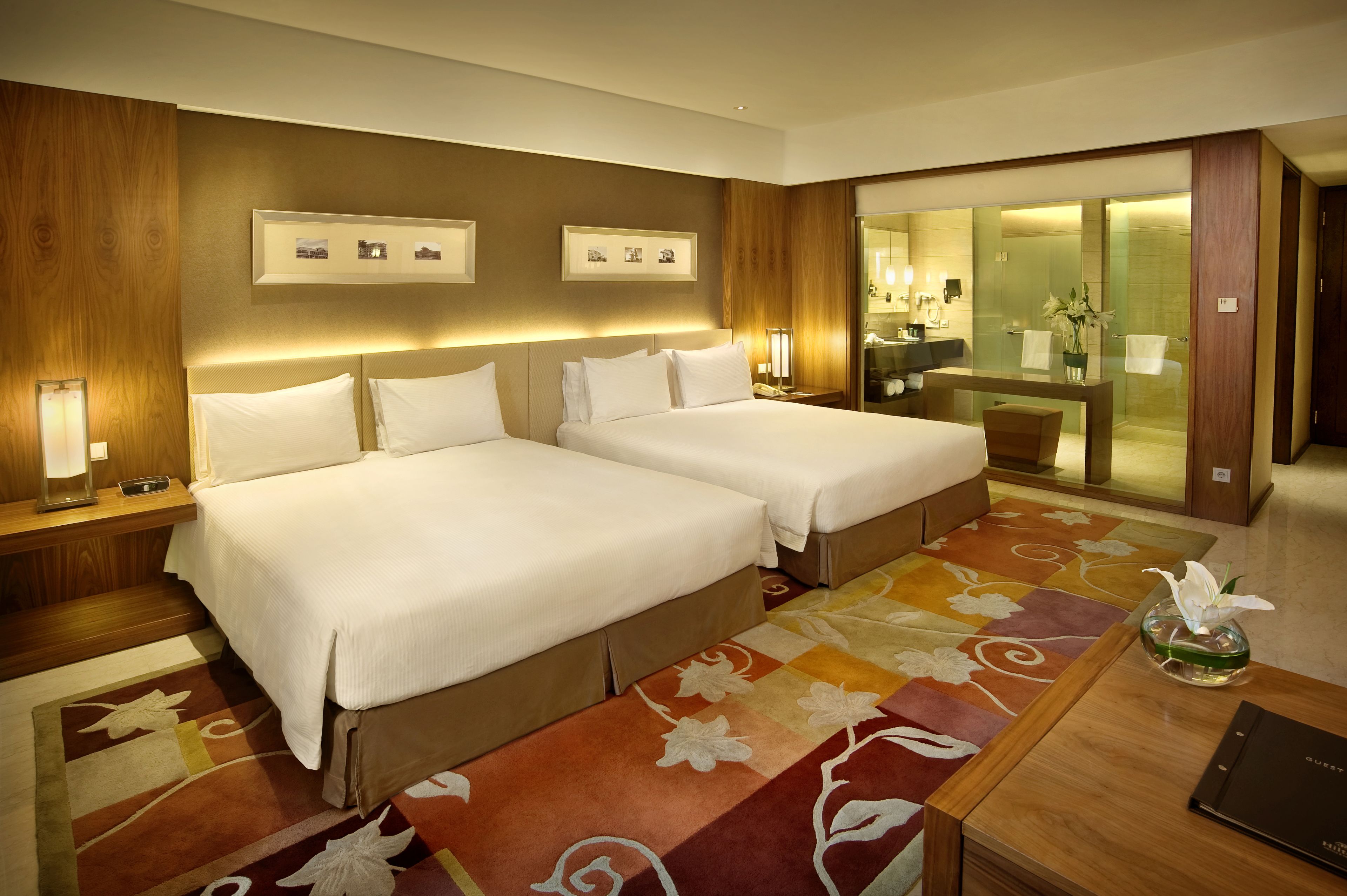 A hotel room with two beds, a large mirror, a bathroom visible through a glass door, and a floral-patterned carpet.