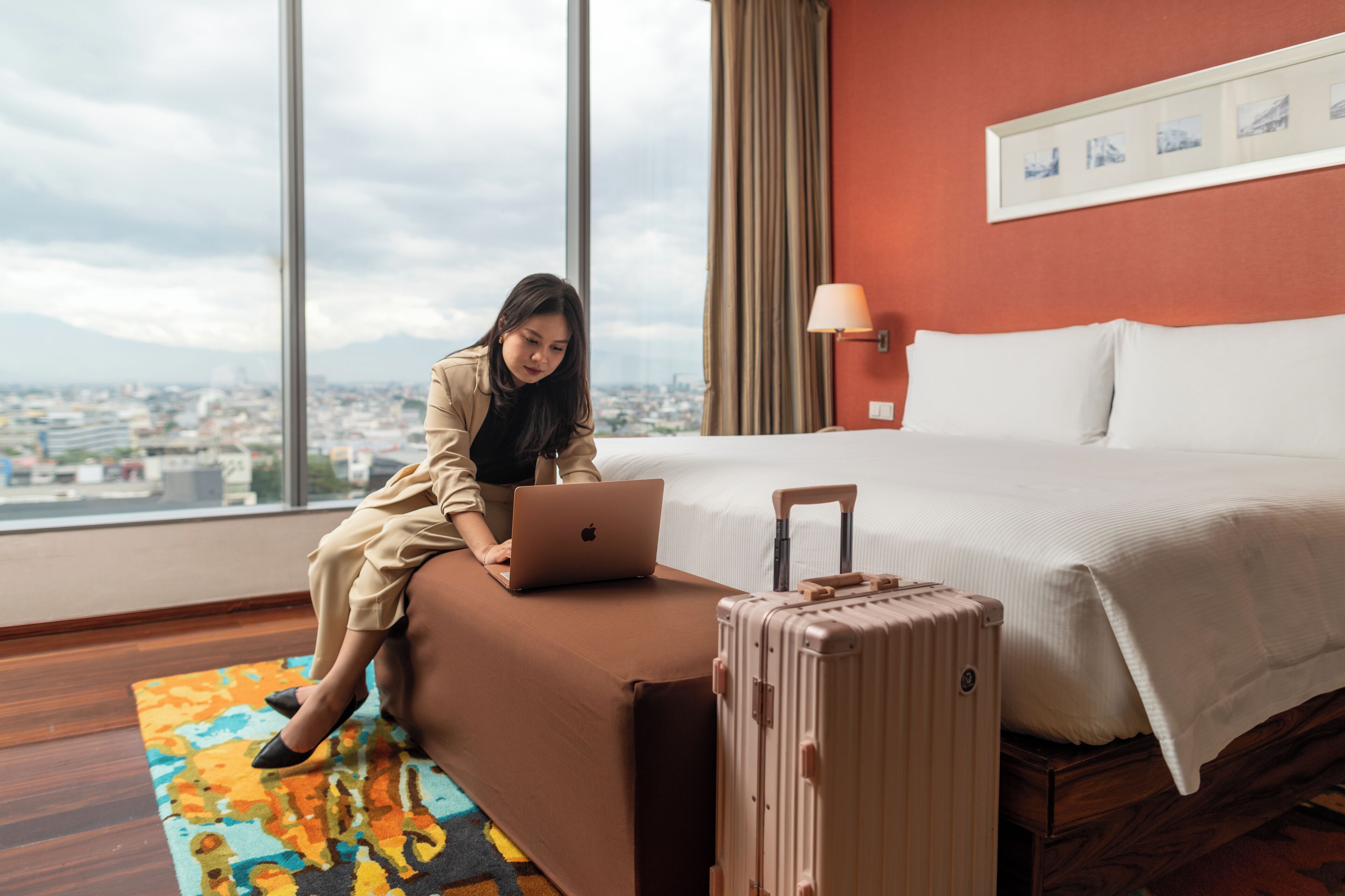 A room with a bed, a window with a city view, a laptop on a seat, and a suitcase.