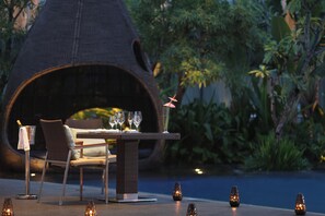 An outdoor dining area with a table set for two, surrounded by chairs, a decorative lamp, and a unique conical structure in the background. - Hilton Bandung (Bandung)
