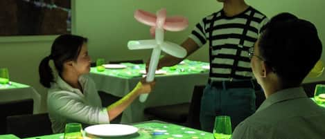 Un espace repas avec des tables préparées pour deux personnes, une personne tenant un ballon et une autre personne debout à côté de la table.