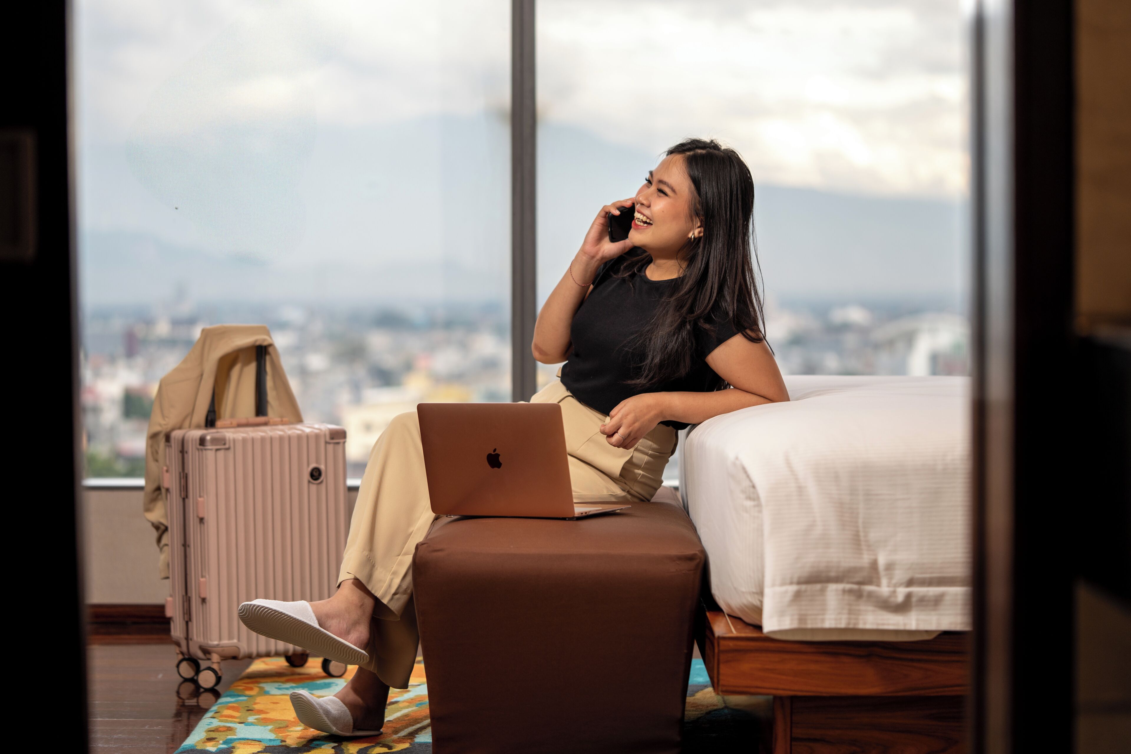 A person sitting on a bed with a laptop, a suitcase, and a bag on the floor.