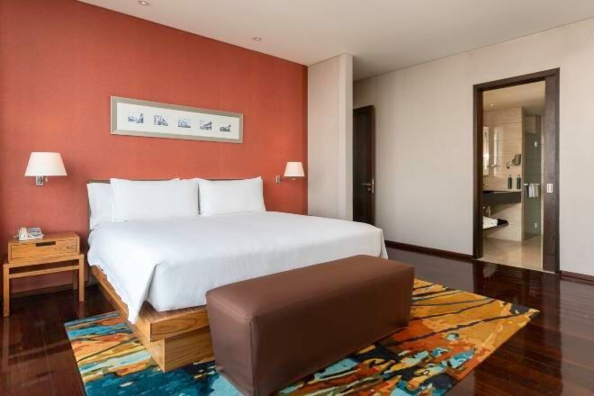 A hotel room with a large bed, a wooden nightstand, a bench, and a colorful rug.