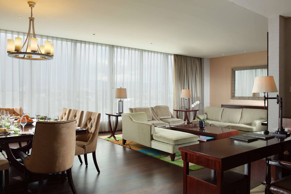 Ett modernt hotellrum med en matplats, två soffor, ett skrivbord och ett stort fönster med tunna, genomskinliga gardiner.