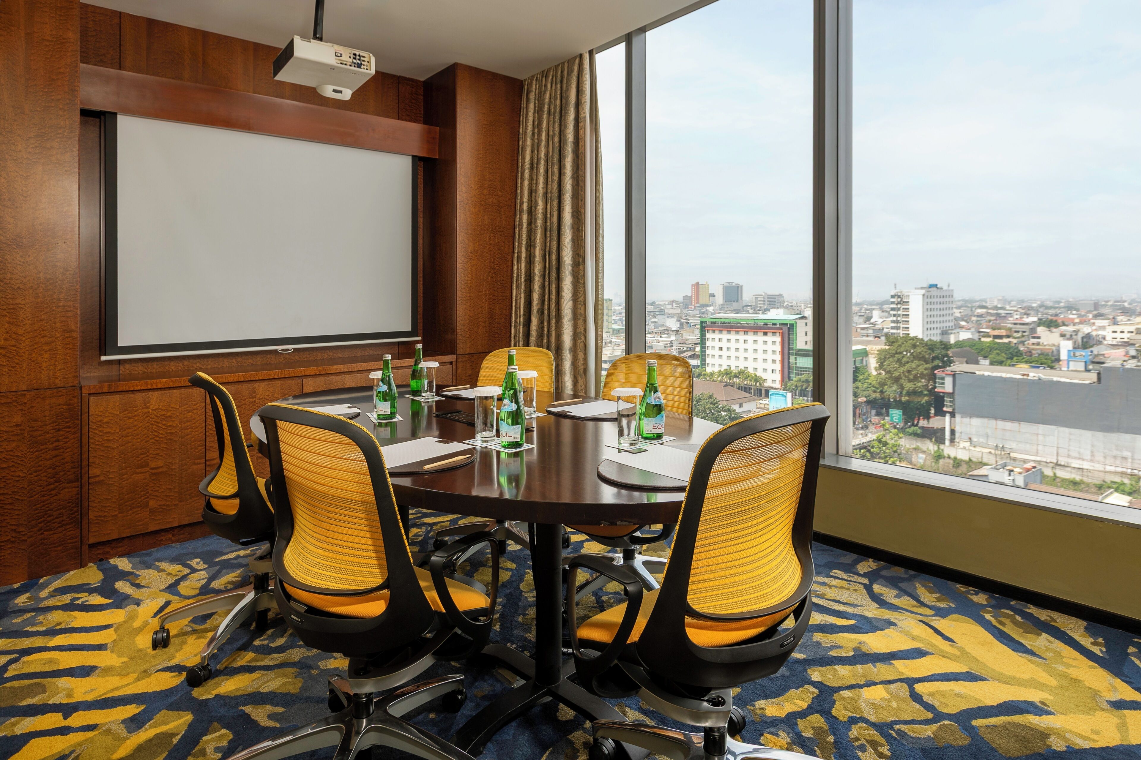 Un salón de conferencias con una gran mesa, cuatro sillas, una pantalla de proyector y vistas a un paisaje urbano a través de amplios ventanales.