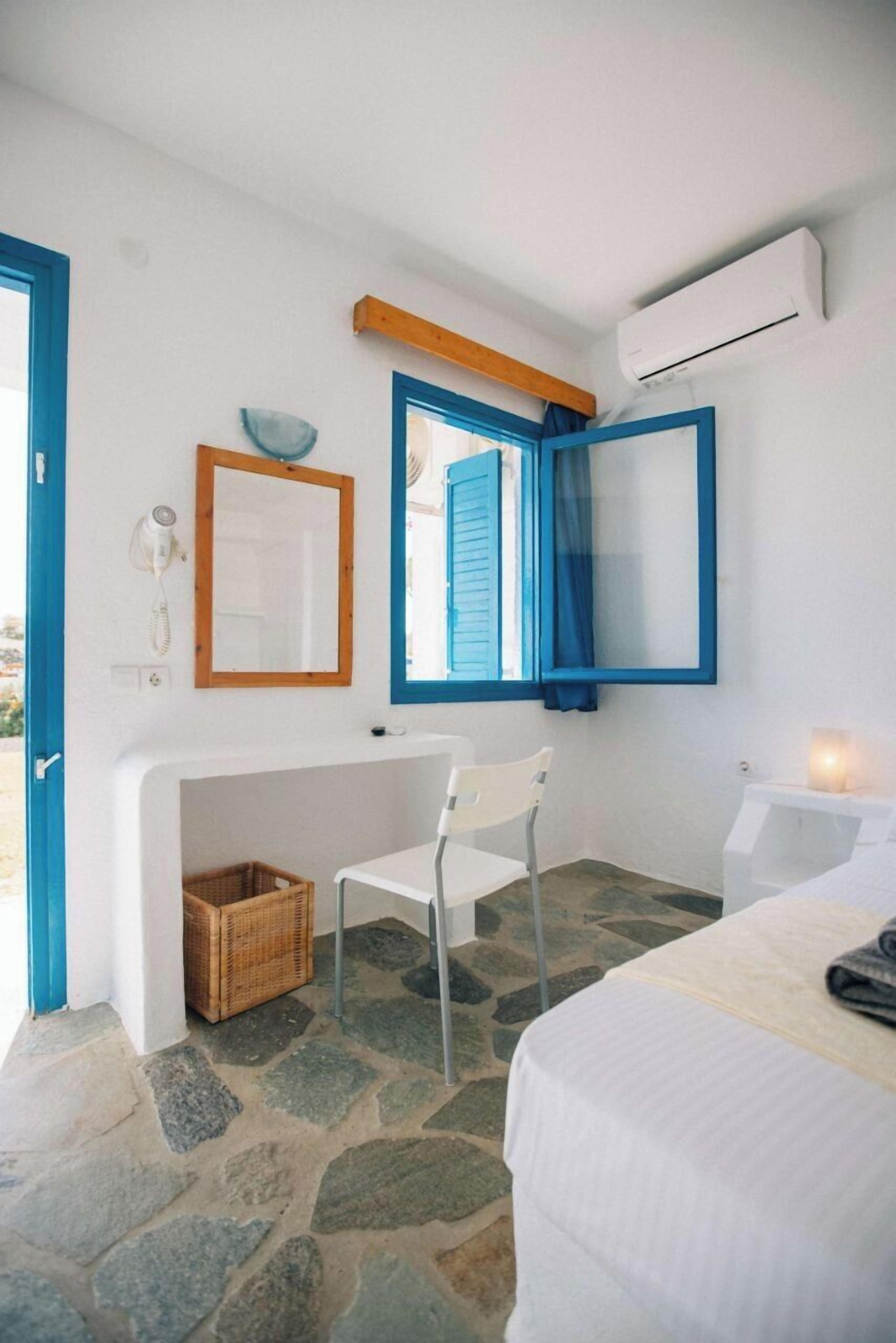 Una habitación con suelo de piedra, una cama blanca, una silla, una mesita con una vela y una ventana con contraventanas azules.