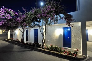 A row of blue doors with white walls, surrounded by flowering plants and trees, under nighttime lighting. - Lagada Beach Hotel (Milos)