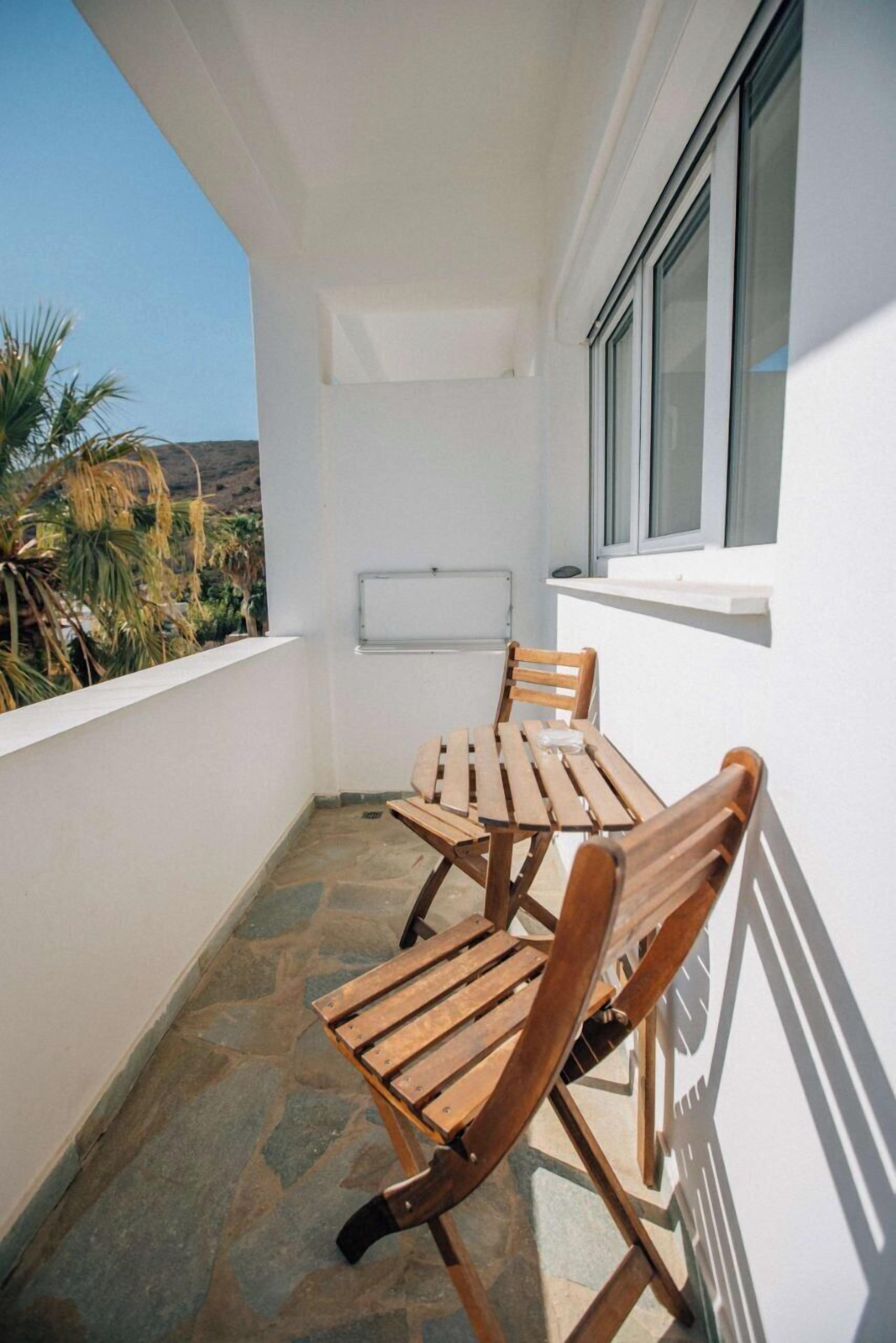 A balcony with a wooden table and chairs, a white wall, and a window.