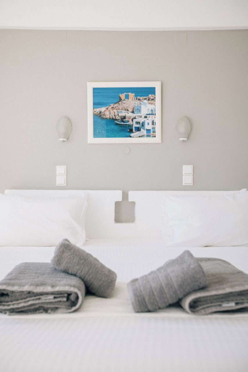 Ein Hotelzimmer mit einem Bett, weißen Kissen, einem gefalteten grauen Handtuch und einem gerahmten Bild einer Küstenlandschaft an der Wand.