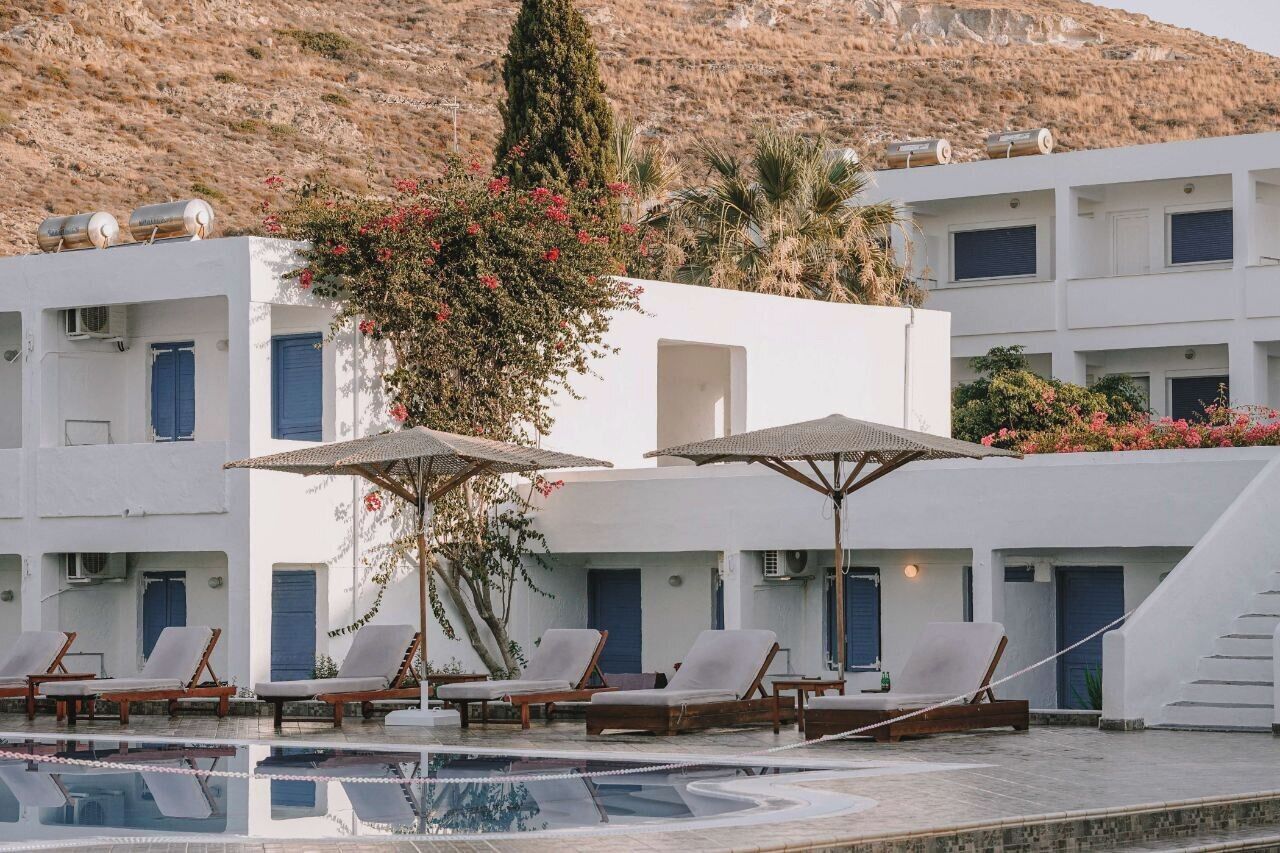 Área de piscina com espreguiçadeiras e guarda-sóis, cercada por edifícios brancos com persianas azuis, emoldurada por um horizonte com encostas.