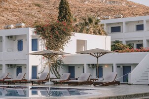 Área de piscina com espreguiçadeiras e guarda-sóis, cercada por edifícios brancos com persianas azuis, emoldurada por um horizonte com encostas.