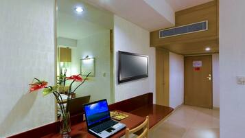 A hotel room with a wooden desk, a laptop, a vase with flowers, a flat-screen TV, and a door with a red sign.