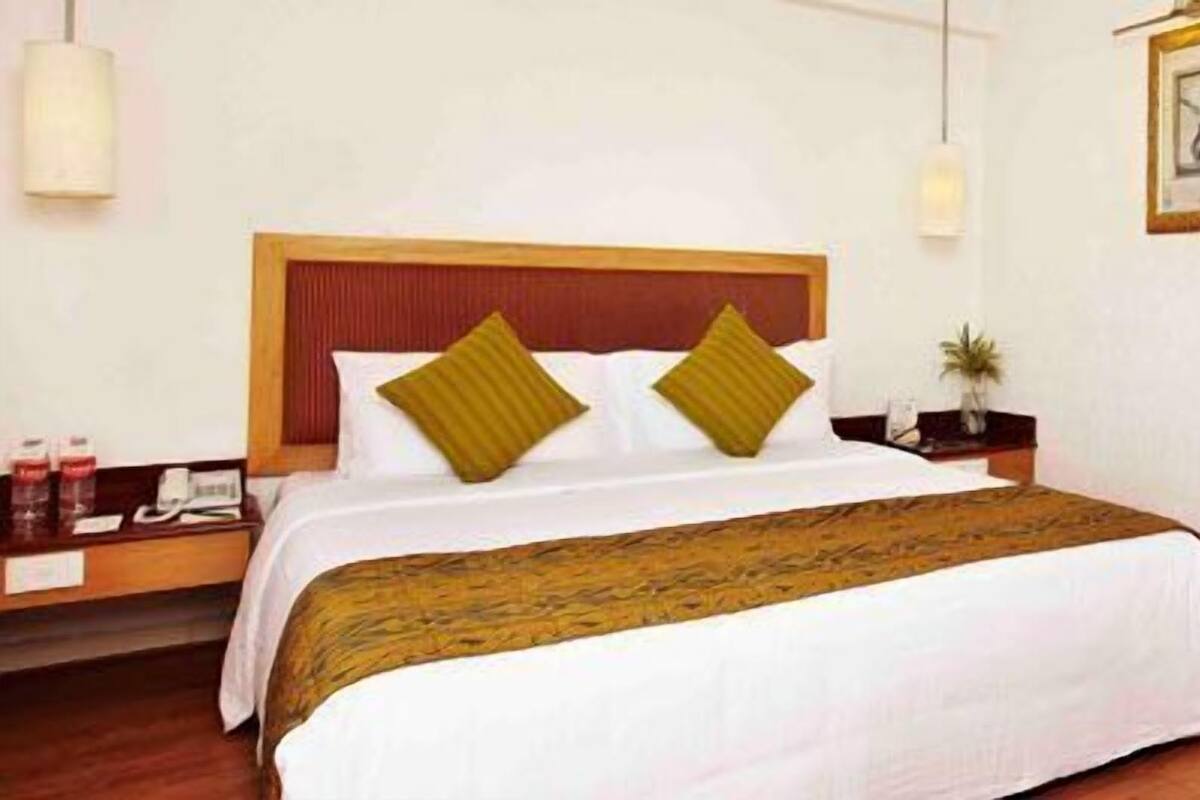 A hotel room with a large bed, wooden headboard, bedside tables, a nightstand, a small table with a plant, and a door leading to another room.