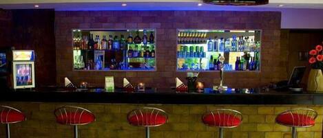 Una zona de bar con dos estantes llenos de botellas de licor, una barra y cuatro taburetes de bar tapizados en rojo.