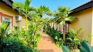 A pathway flanked by lush greenery and yellow buildings with red roofs, leading to a covered area.