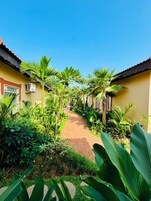 A pathway flanked by lush greenery and yellow buildings with red roofs, leading to a covered area.