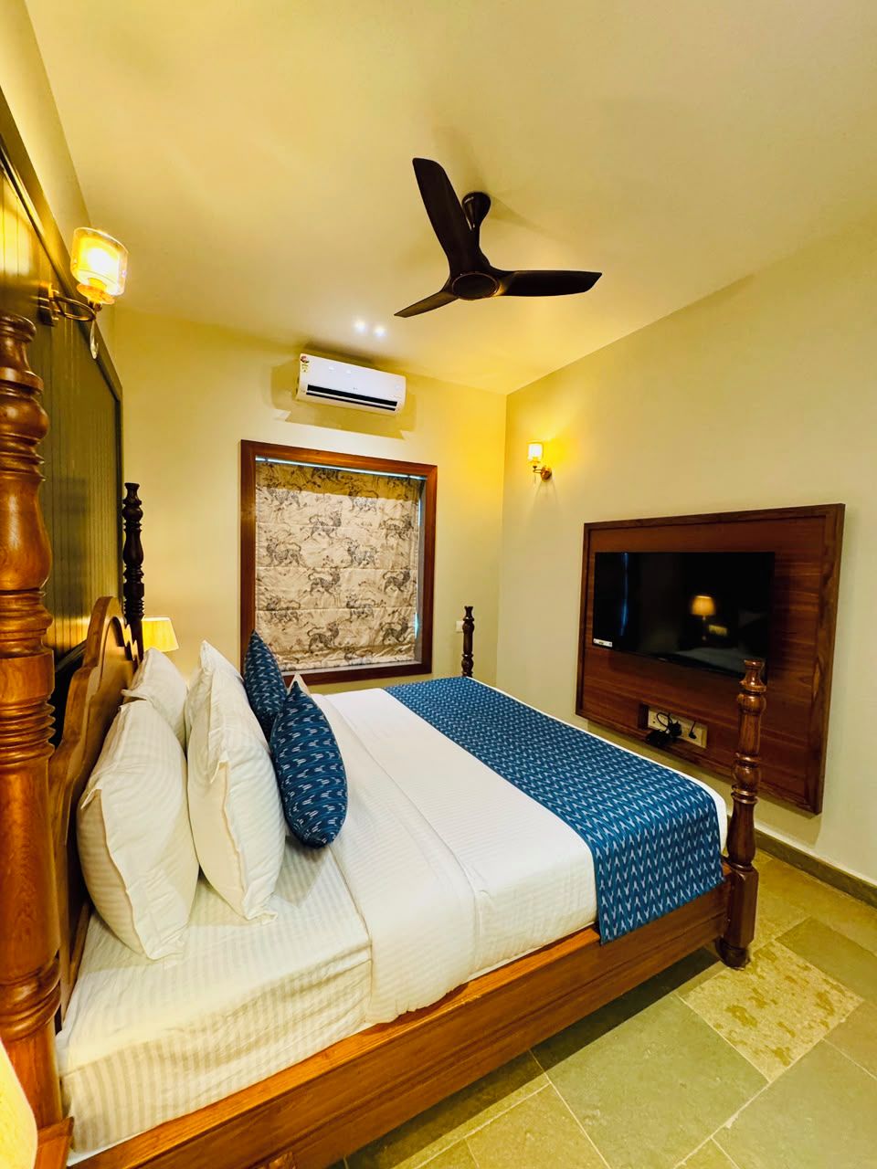 A bedroom with a bed, a ceiling fan, an air conditioning unit, a television mounted on the wall, and a decorative wall hanging.