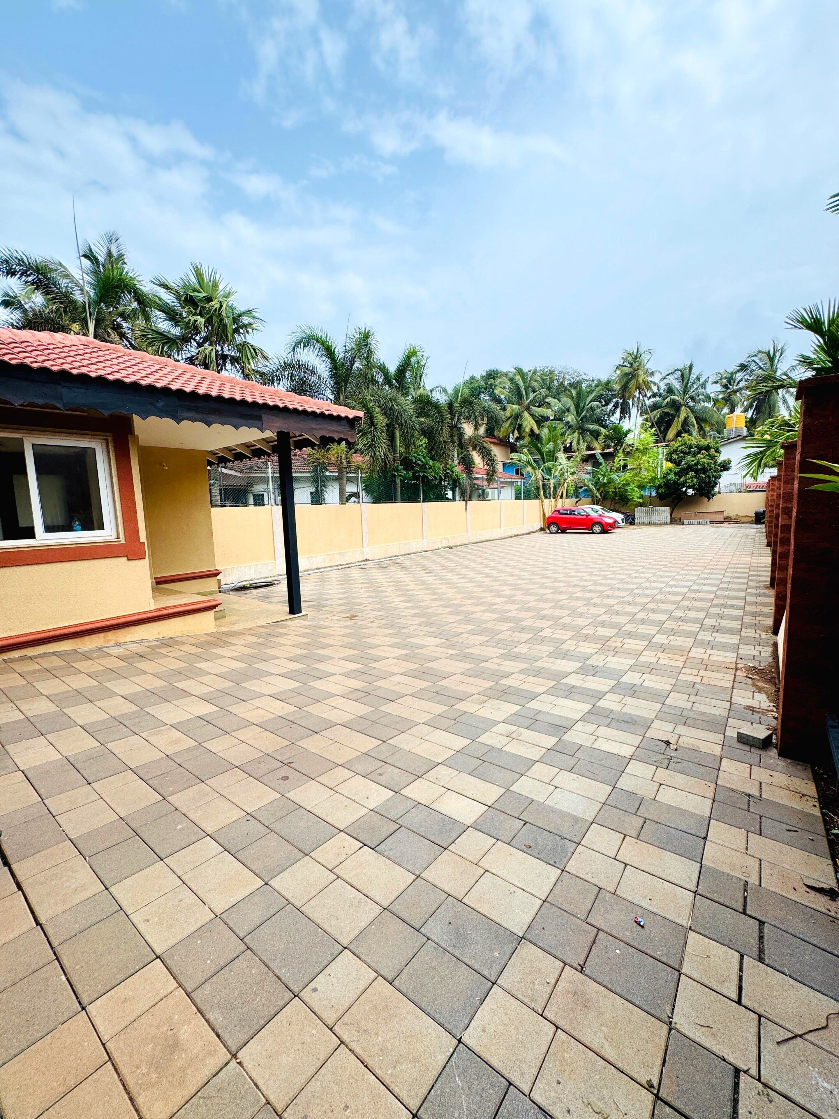 A paved courtyard with a tiled floor, a small building with a red roof, and a red car parked in the distance.