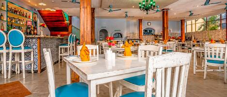 Un restaurante con mesas blancas y sillas azules, una zona de bar con estantes llenos de botellas y una barra roja.