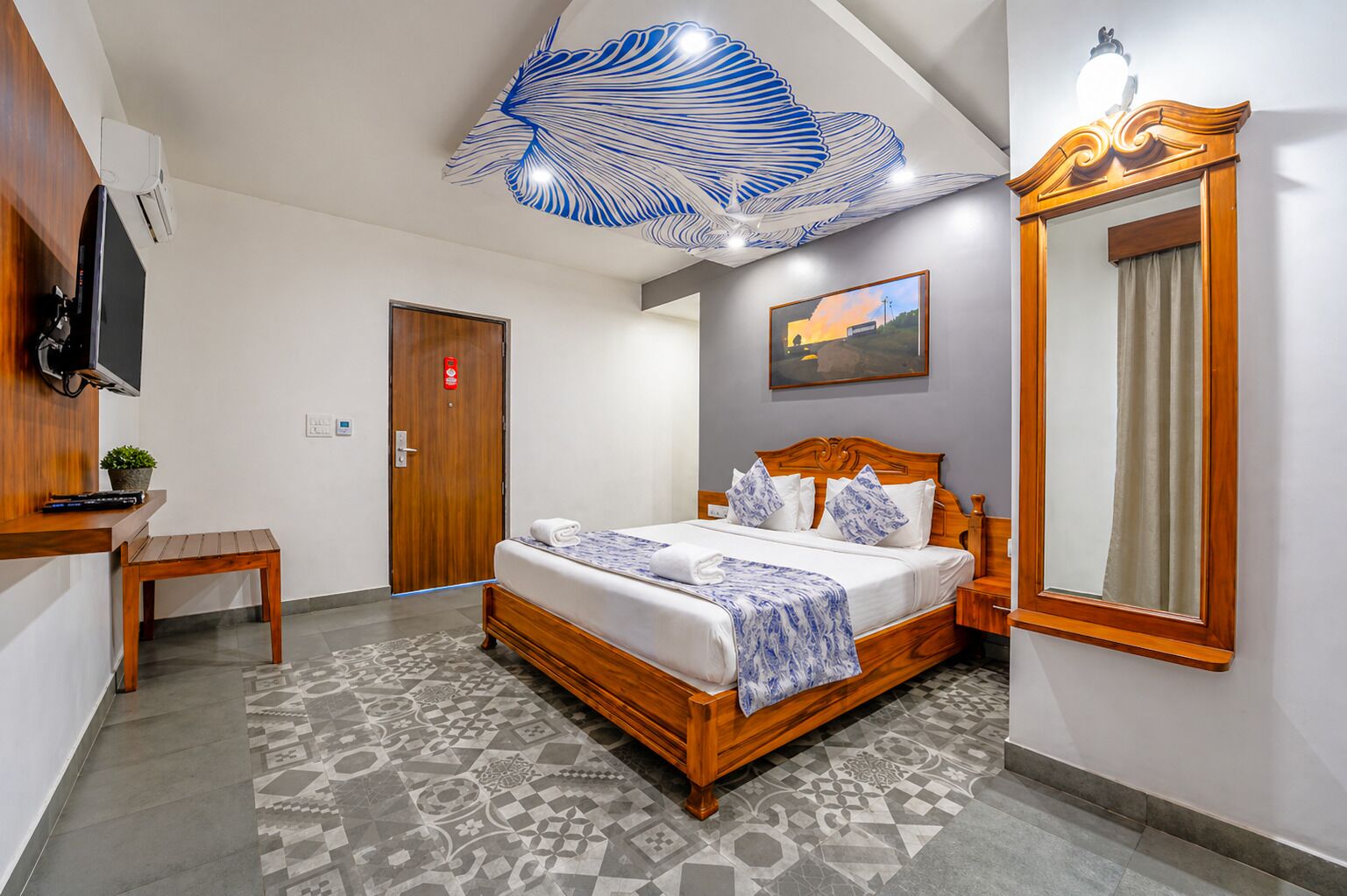 A hotel room with a bed, a desk, a chair, a mirror, and a ceiling light fixture.