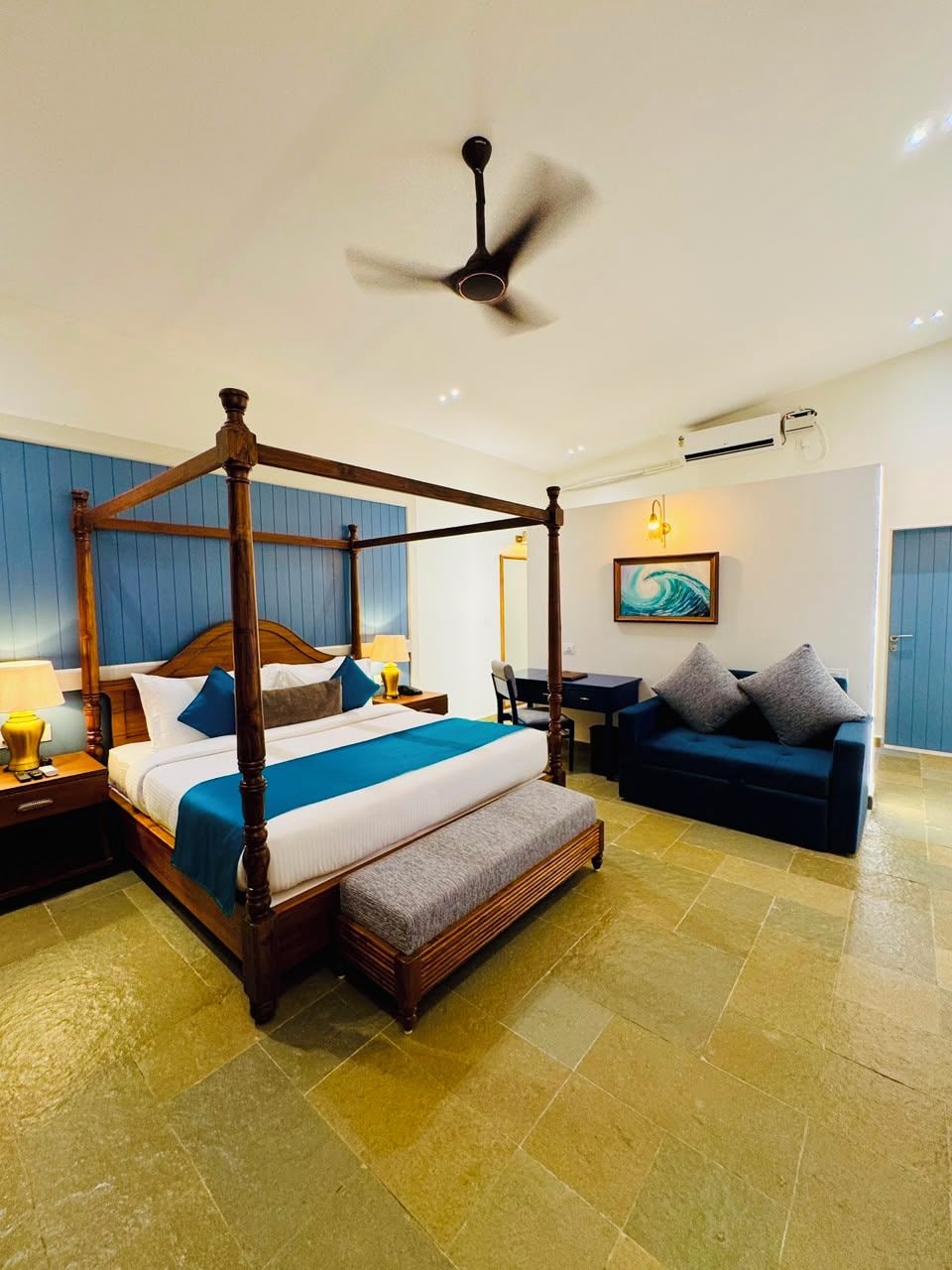 A four-poster bed with a wooden frame, a desk with a chair, a blue sofa, and a ceiling fan.