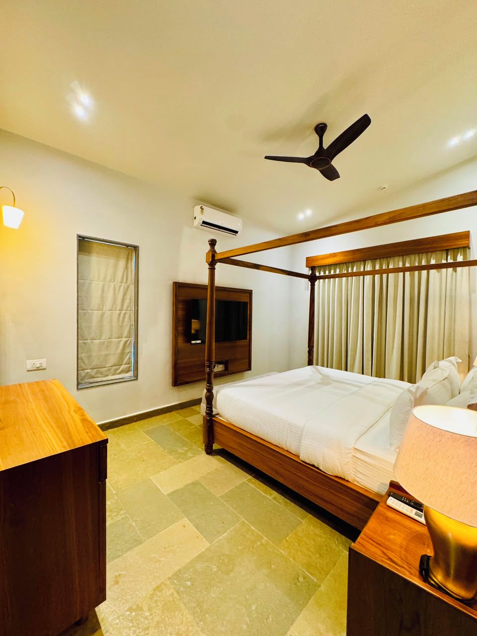 A hotel room with a four-poster bed, a ceiling fan, a TV mounted on the wall, and a bedside table with a lamp.