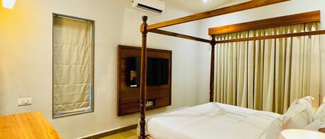 A hotel room with a four-poster bed, a ceiling fan, a TV mounted on the wall, and a bedside table with a lamp.