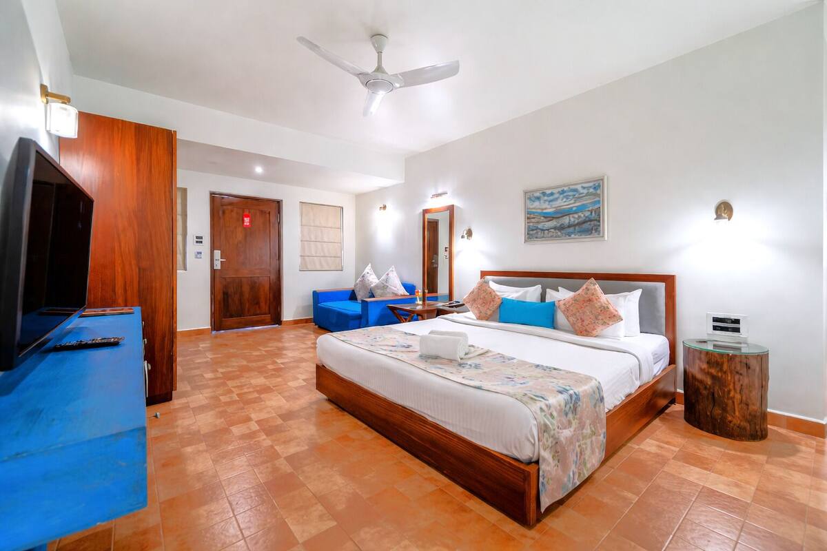 Quarto de hotel com cama, televisão, escrivaninha, cadeira, sofá azul e um quadro na parede.