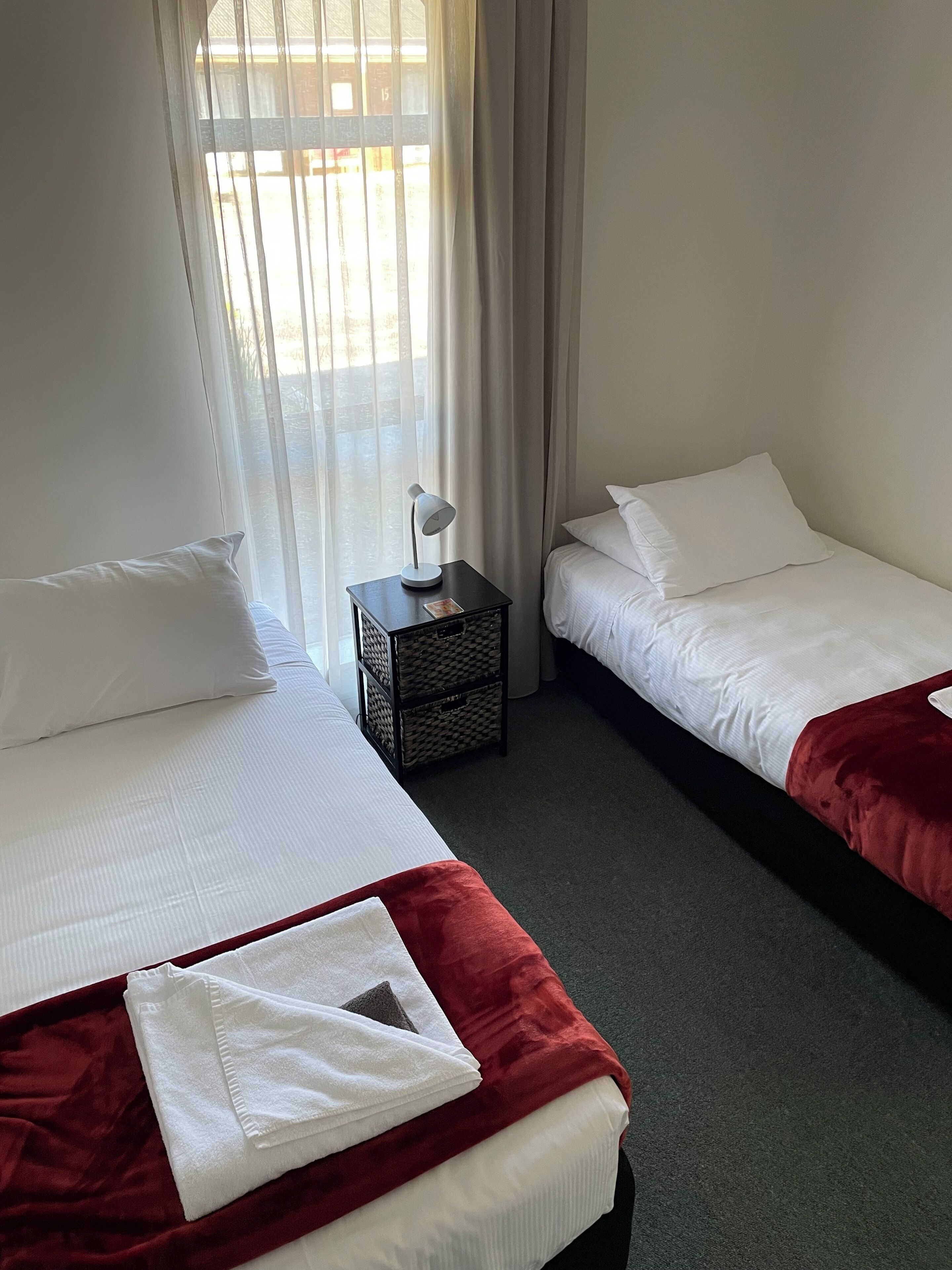 Habitación de hotel con dos camas, una mesita de noche y una ventana con cortinas traslúcidas.