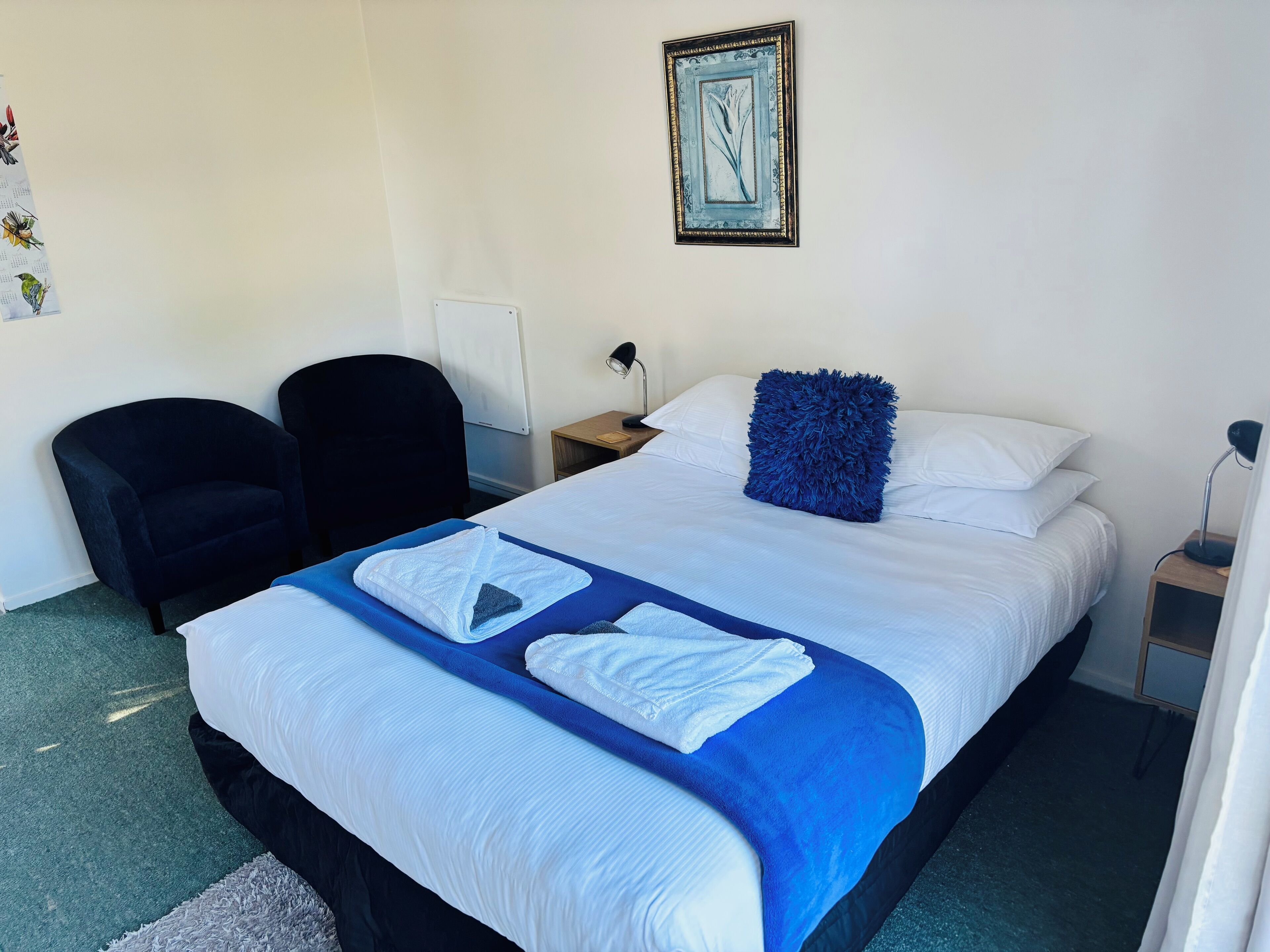 A neatly made bed with white and blue bedding, a blue throw blanket, and a fluffy blue pillow. There is a black chair and a small bedside table with a lamp.