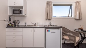 A compact kitchen with white cabinets, a microwave, a sink, and a small refrigerator. There is a window with curtains, a chair, and a table with a candle and a jar. - Buller Bridge Motel (Westport)