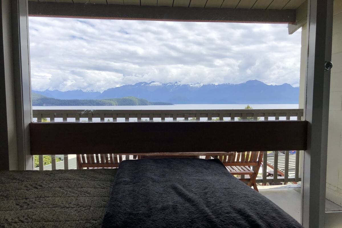 A room with a bed, a view of mountains and a lake through a window.