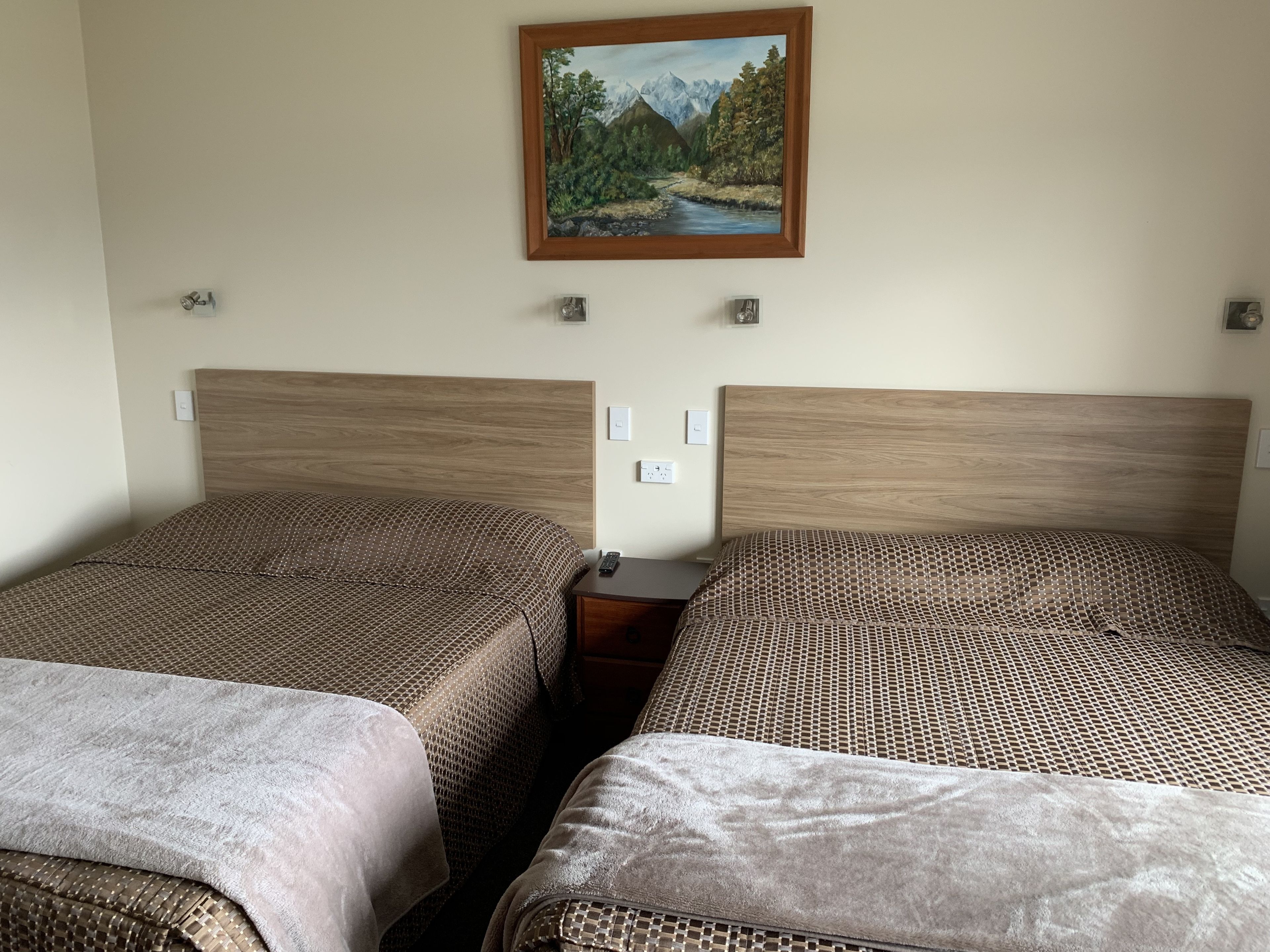 A hotel room with two beds, each with a patterned bedspread, a wooden headboard, and a painting of a mountain landscape on the wall.