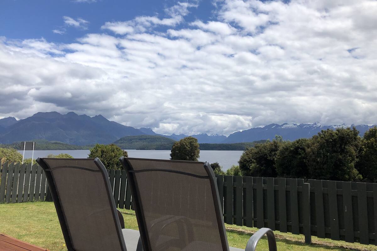A view of a lake with mountains in the background, a wooden fence, and two chairs facing the lake.